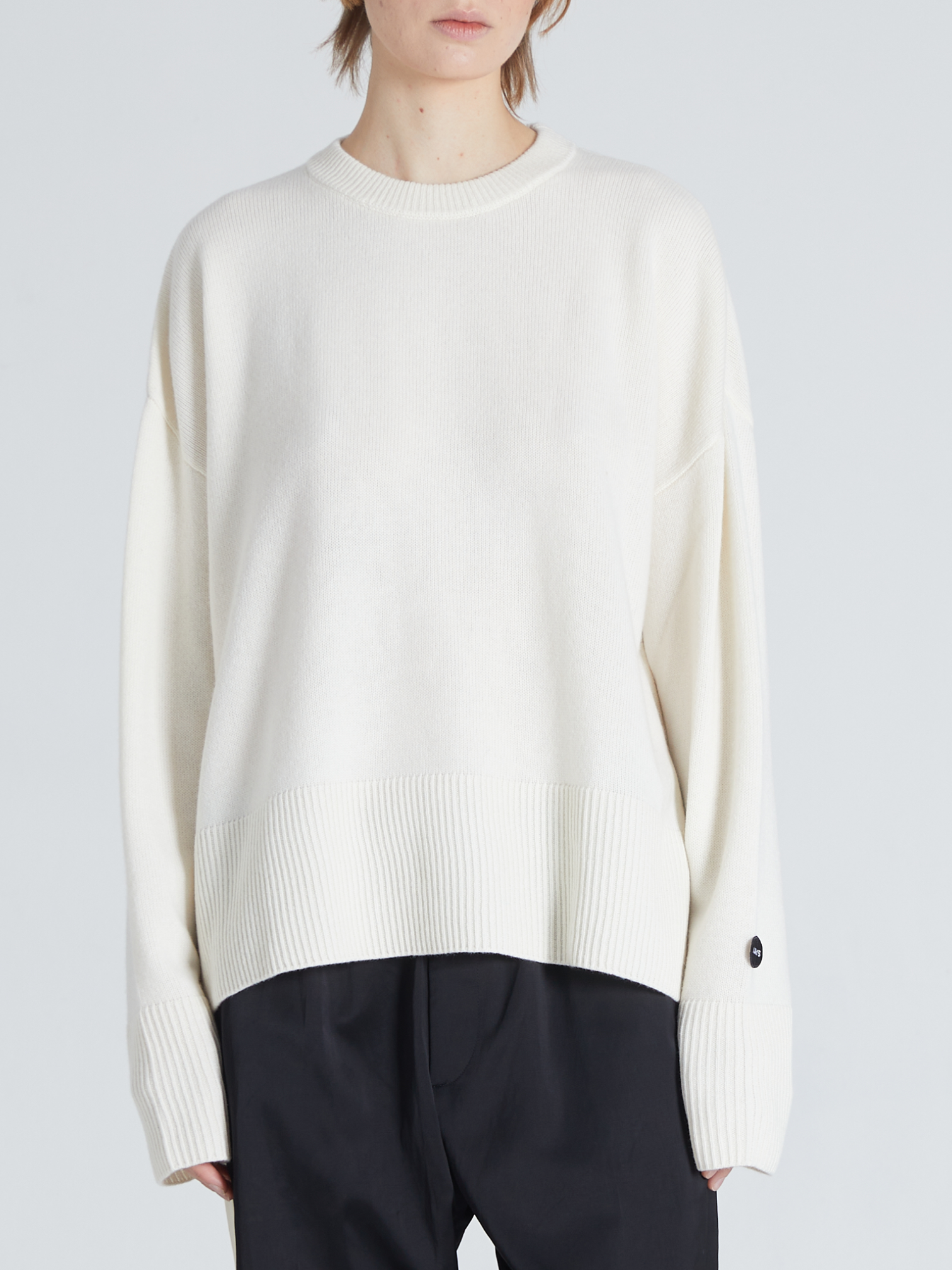 A model wearing a cream-colored knit crewneck sweater with dropped shoulders and ribbed hem/cuffs, paired with dark trousers, against a light gray studio background.