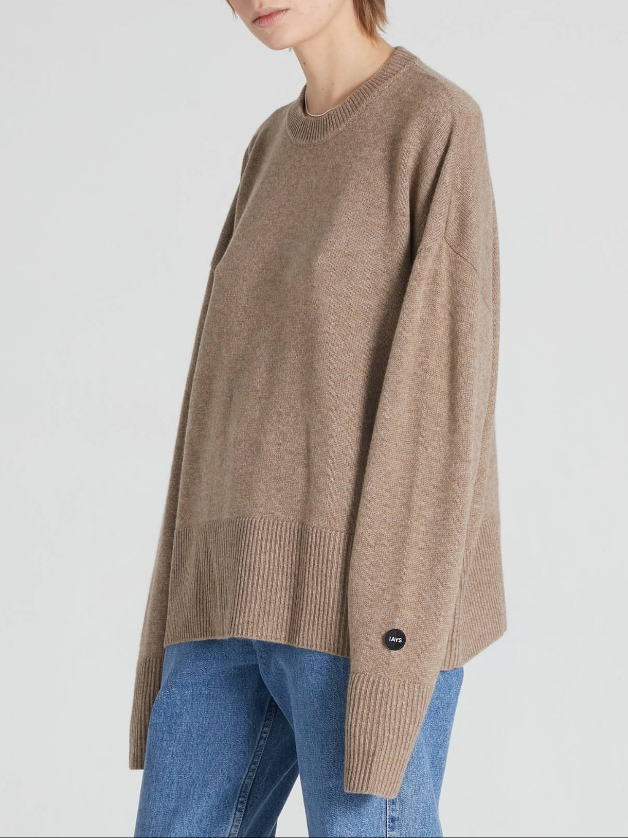 A model wearing an oversized, light brown crewneck knit sweater with ribbed hem and cuffs, paired with blue jeans, against a plain light gray background.