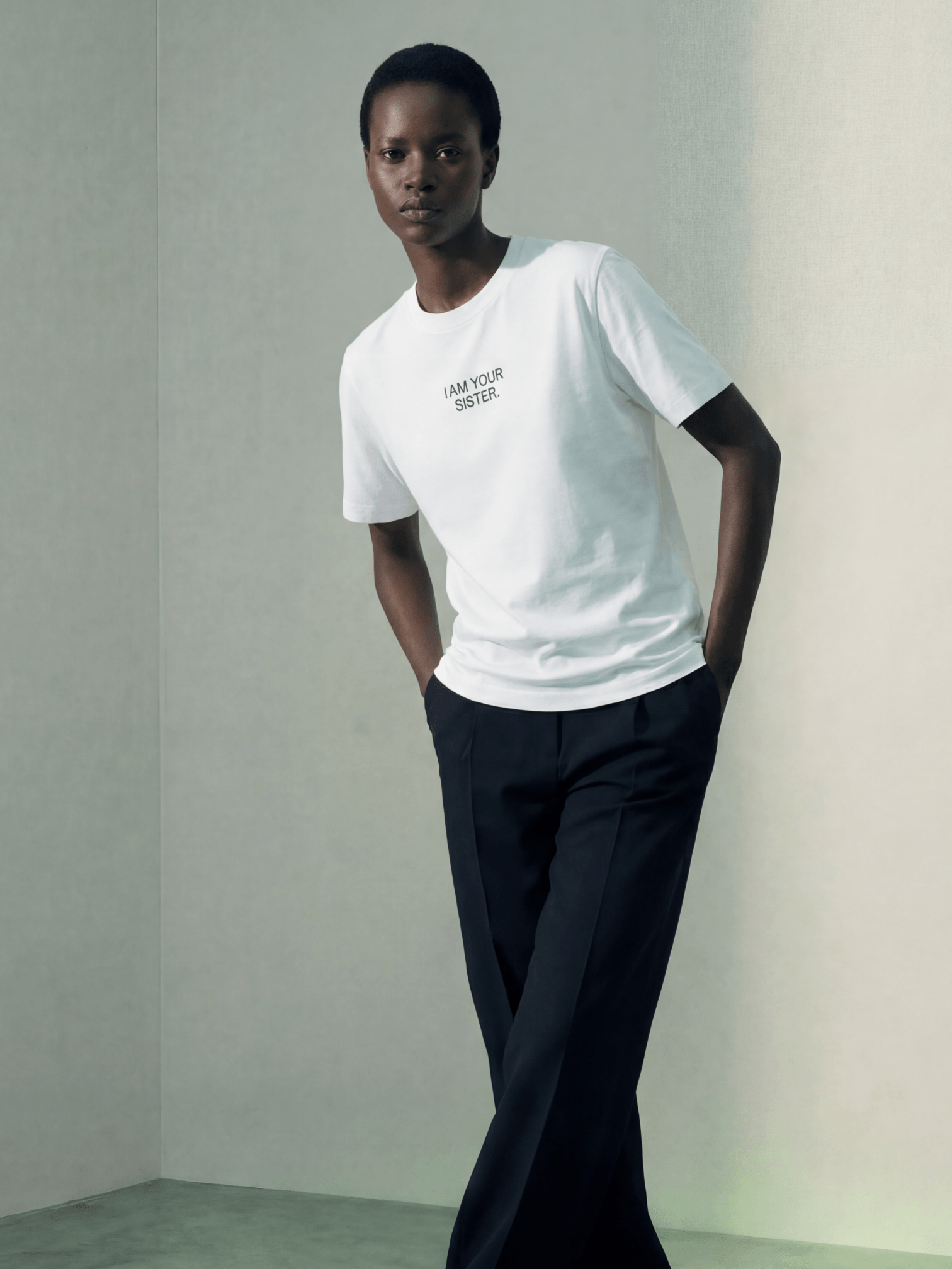 Woman wearing a white Sister t-shirt with text and dark pants against a plain background
