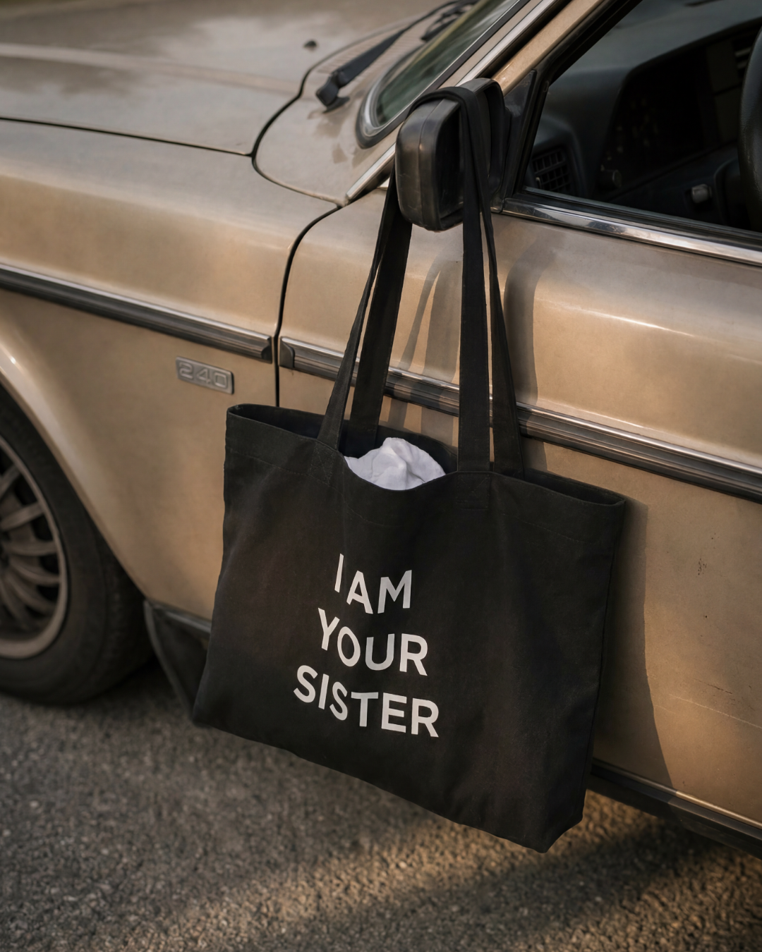Black tote bag with 'I AM YOUR SISTER' text hanging from a car door.
