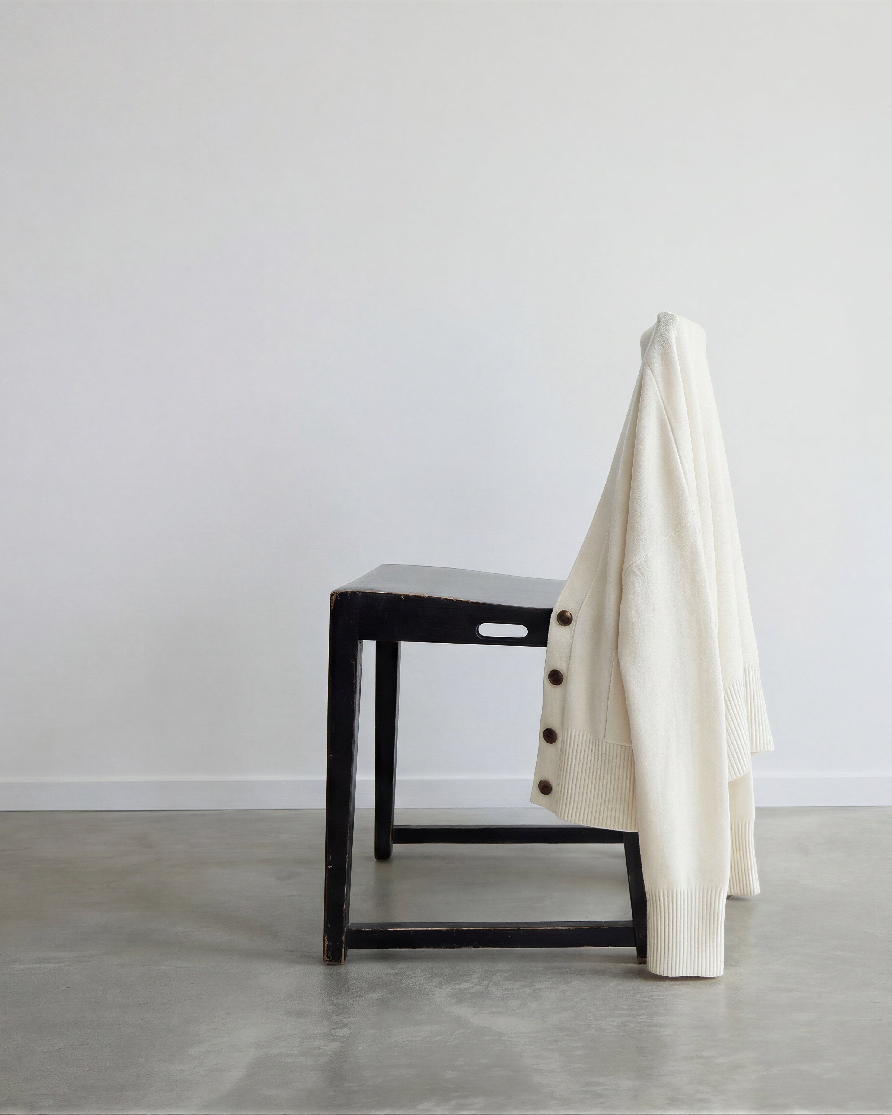 White cashmere cardigan draped over a black chair in a minimalistic room with concrete floor and large windows.