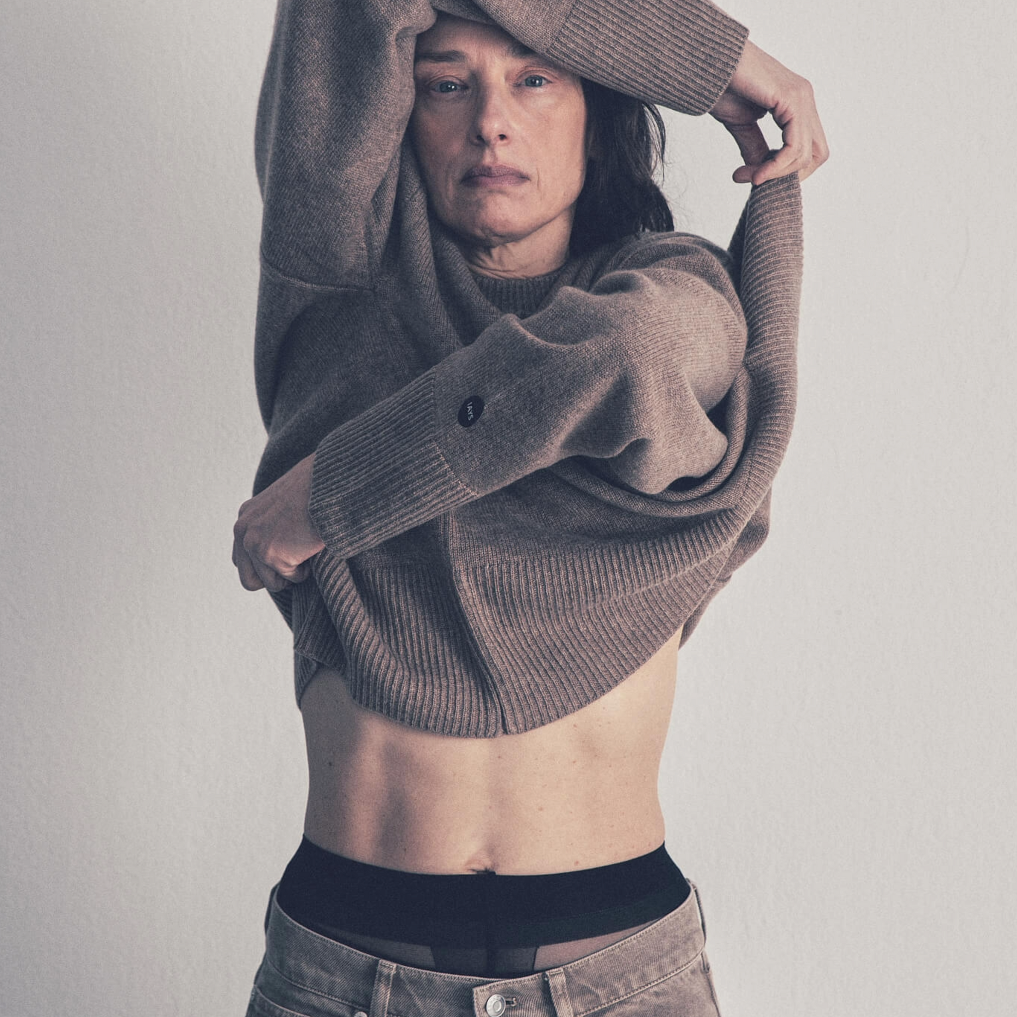 A woman wearing a beige cashmere crew neck sweater with ribbed trims, paired with beige trousers. The sweater has a relaxed fit with a classic rounded neckline.