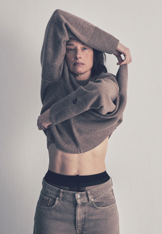 A woman wearing a beige cashmere crew neck sweater with ribbed trims, paired with beige trousers. The sweater has a relaxed fit with a classic rounded neckline.