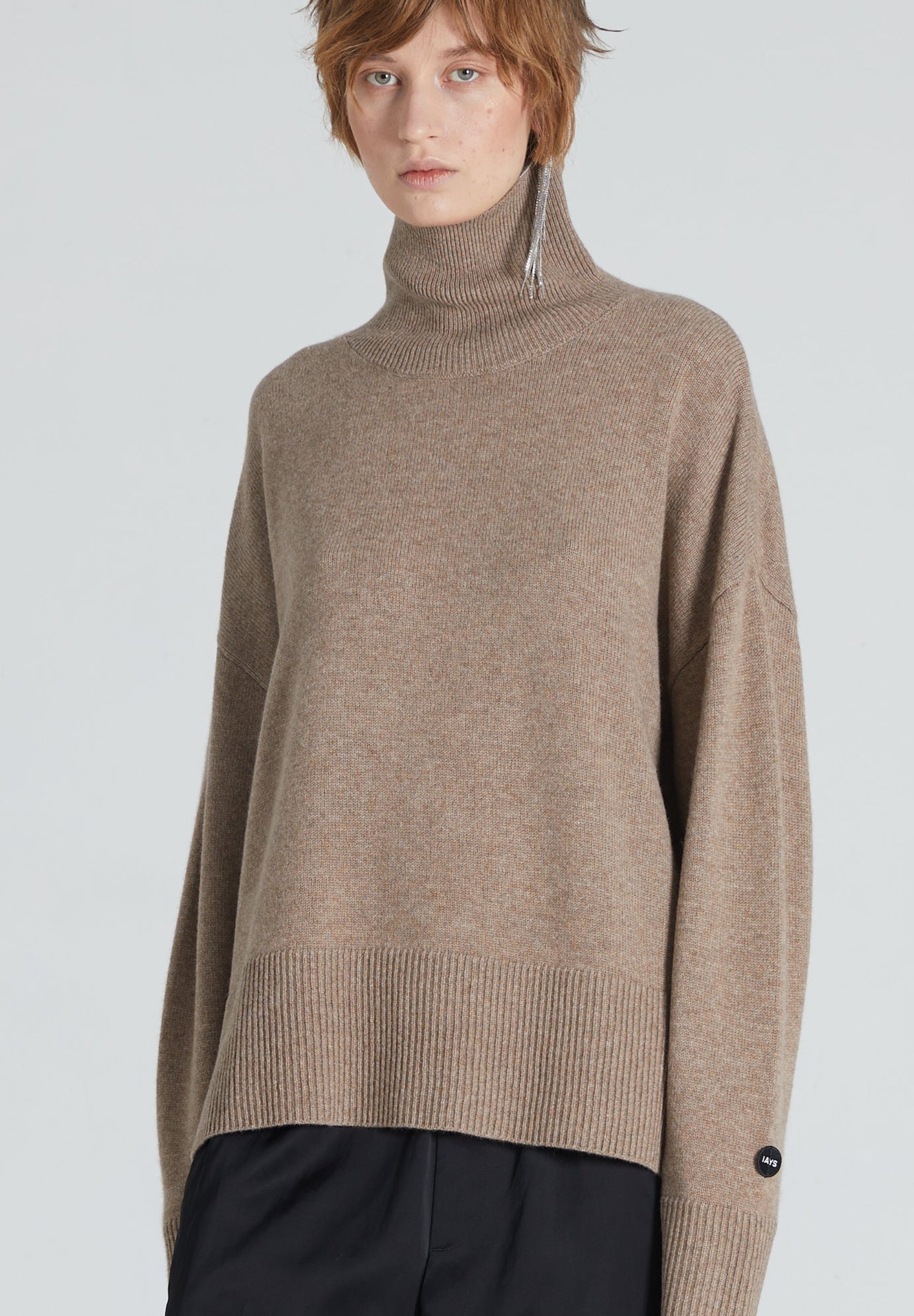 A model wearing a light brown knit turtleneck sweater with a high ribbed hem and cuffs, paired with black trousers, against a solid light gray background.
