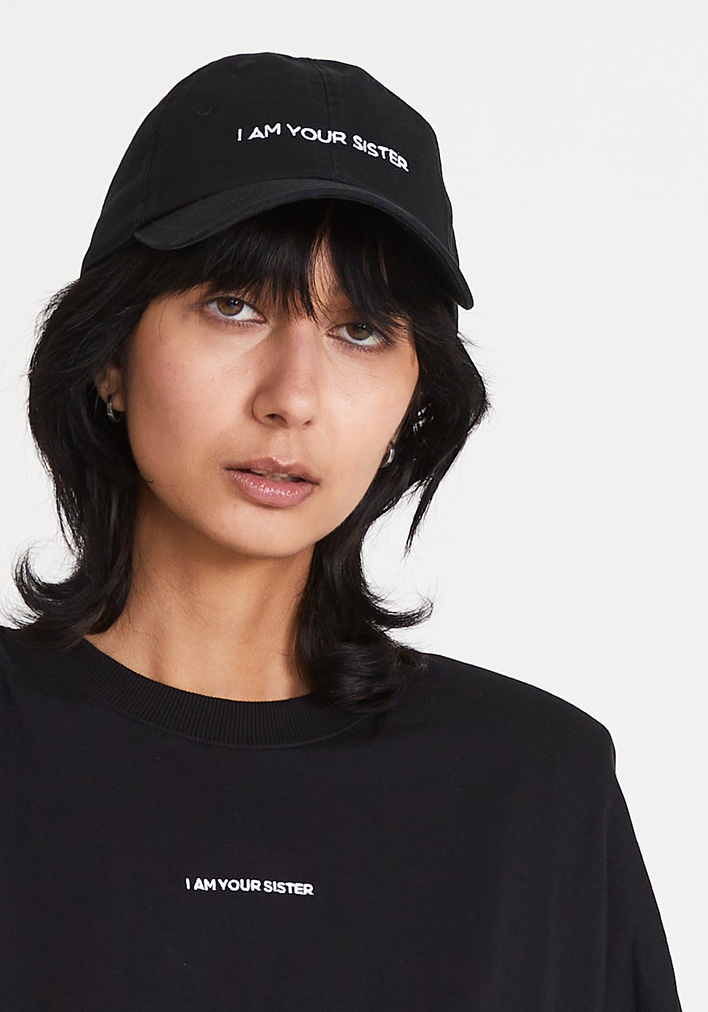 A woman wearing a black baseball cap with an embroidered 'I AM YOUR SISTER' logo on the front.