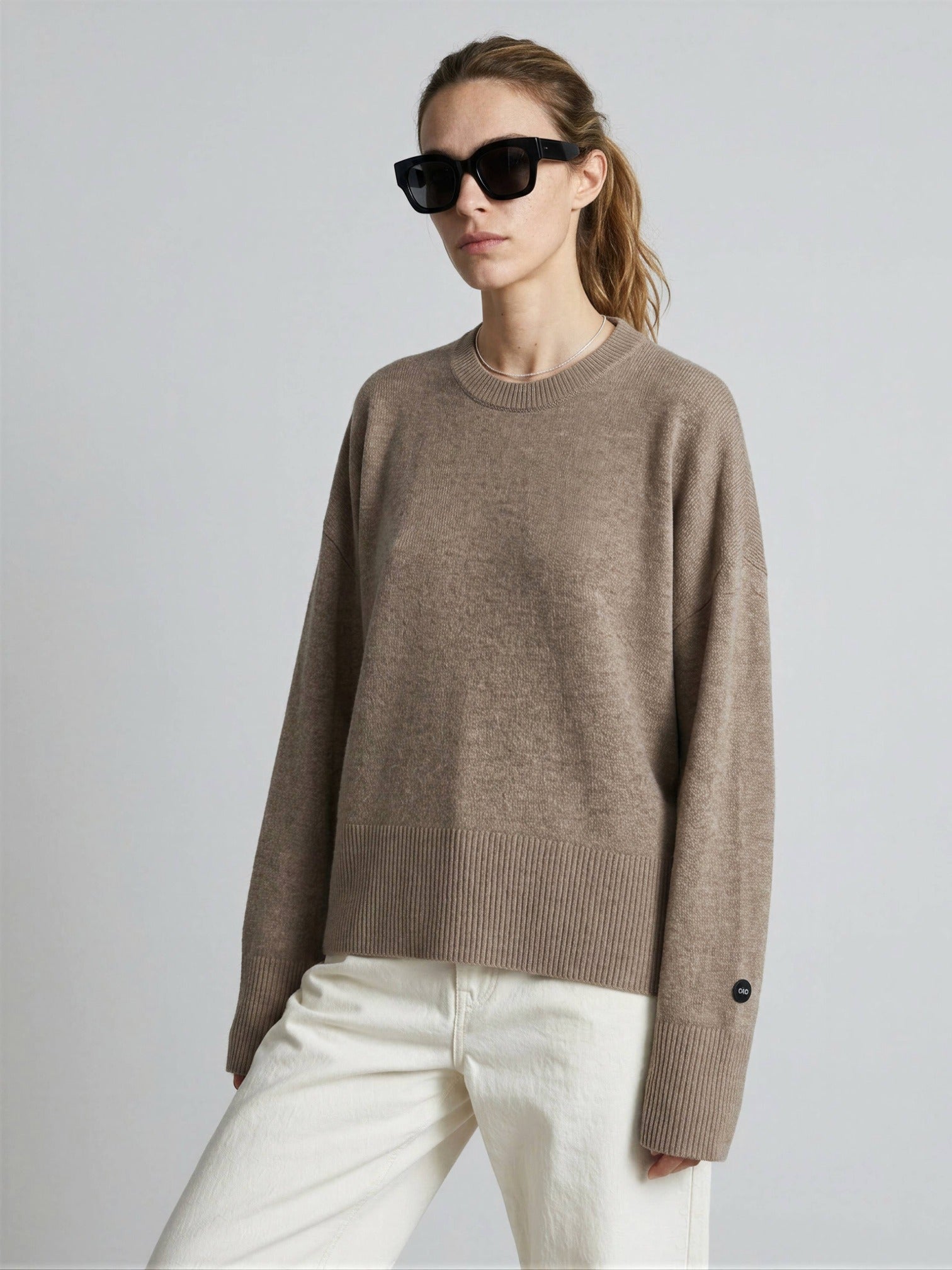 Woman wearing a brown cashmere sweater and white pants with sunglasses on a plain background