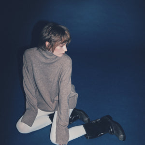 A woman wearing a brown cashmere turtleneck sweater with ribbed trims, featuring a standing neck, dropped shoulders, and extended sleeves, is sitting against a blue background.