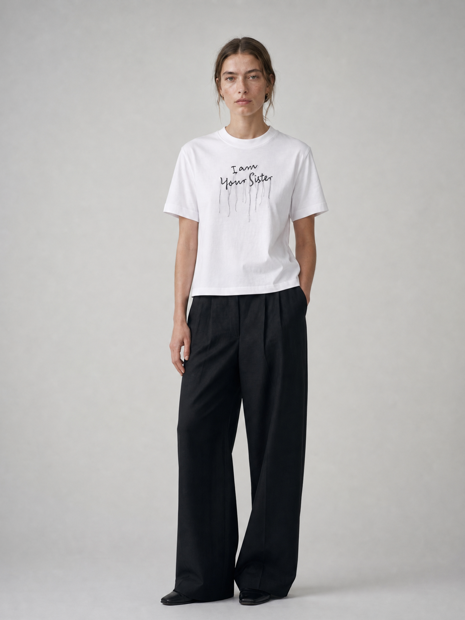 A model stands against a light gray studio background wearing a white I am your sister T-shirt with black embroidered text and black wide-leg trousers.