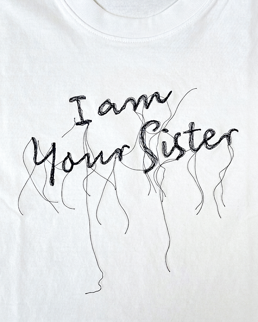 A close-up of a white t-shirt featuring the phrase 'I am Your Sister' embroidered in a black, hand-drawn cursive font with messy, dangling line details.